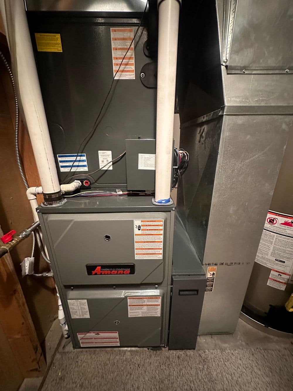 Amana furnace installation in a residential utility room with labels and pipes visible.