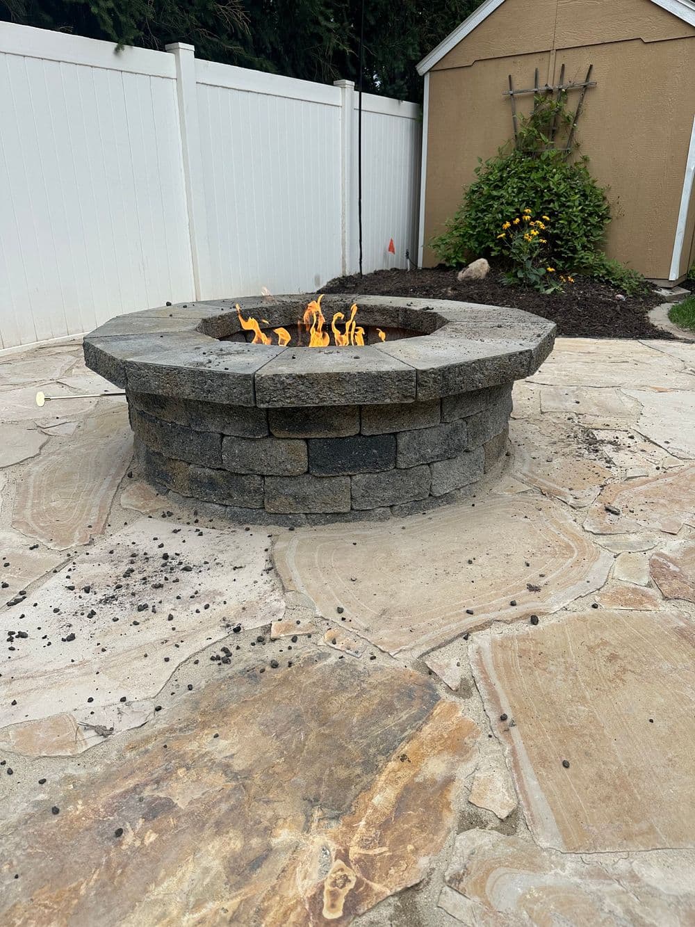 Stone fire pit surrounded by a paved patio and white fence, featuring a vibrant flame.