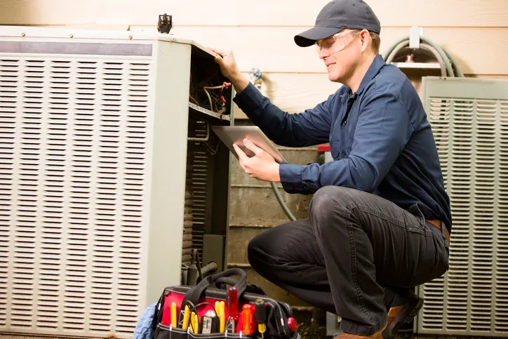 Man with a baseball cap crouching and working on an outdoor HVAC unit.