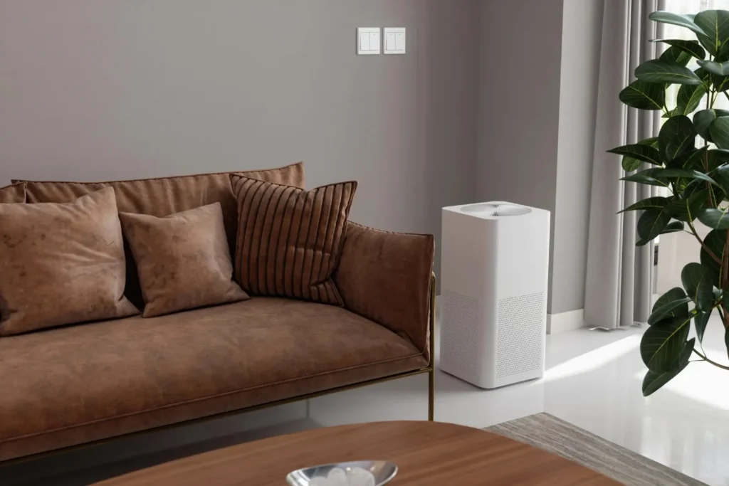 A white whole home air dehumidifier stands next to a brown velvet sofa with throw pillows in a modern living room, with a large green plant and a window with sheer curtains visible to the right.