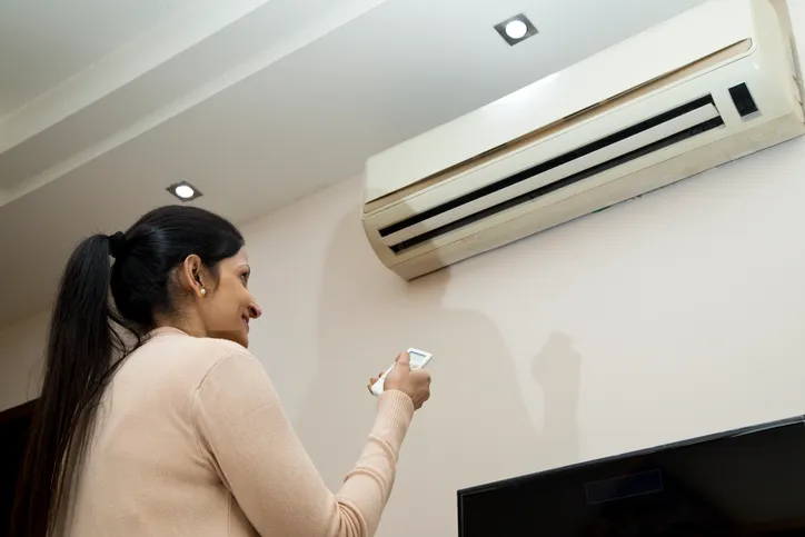 A woman smiling and holding a remote control pointed at a wall-mounted mini-split air conditioner.