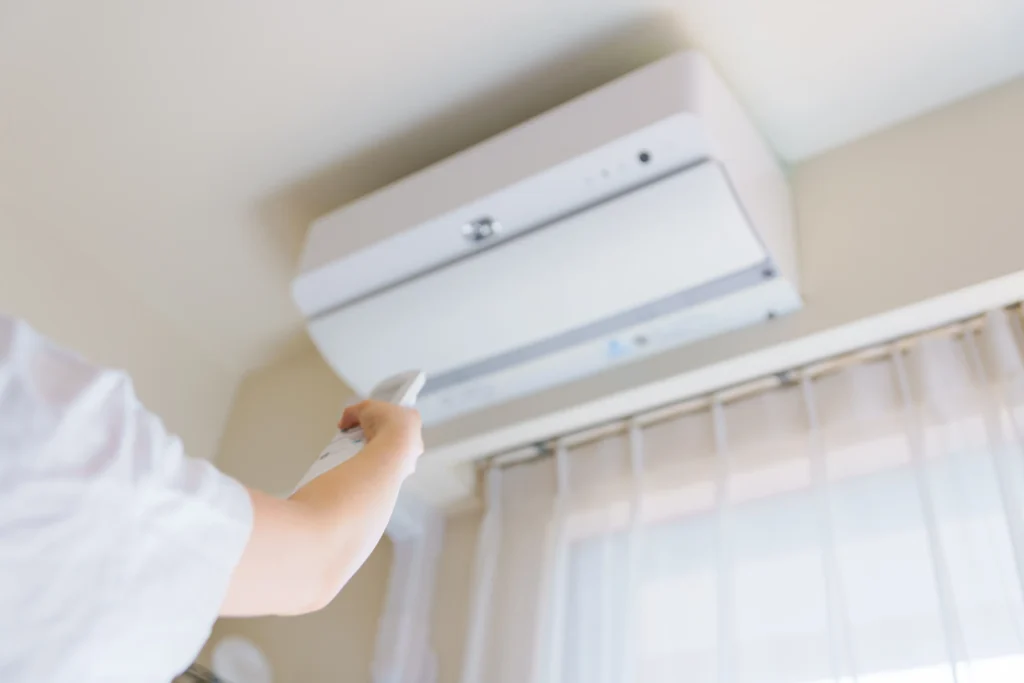 Person using remote control to operate white wall-mounted air conditioning unit above window with sheer curtains